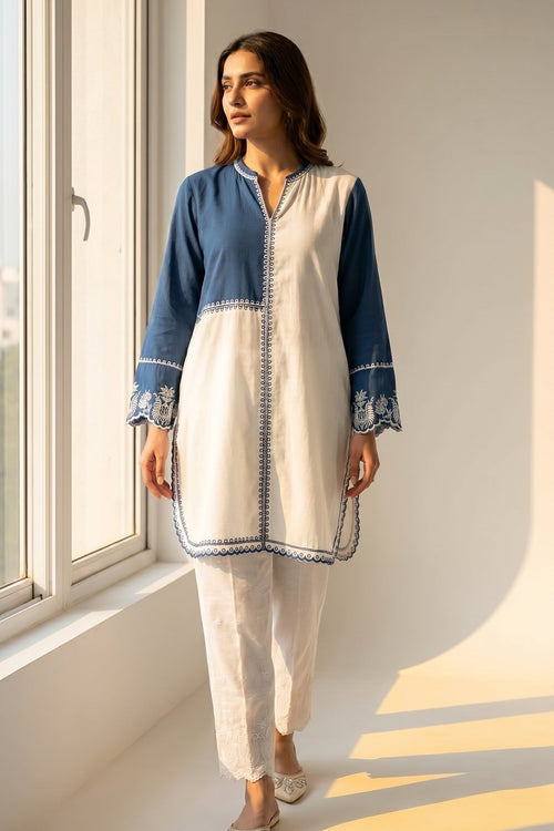 Woman wearing a blue and white traditional outfit standing by a window with sunlight streaming in.